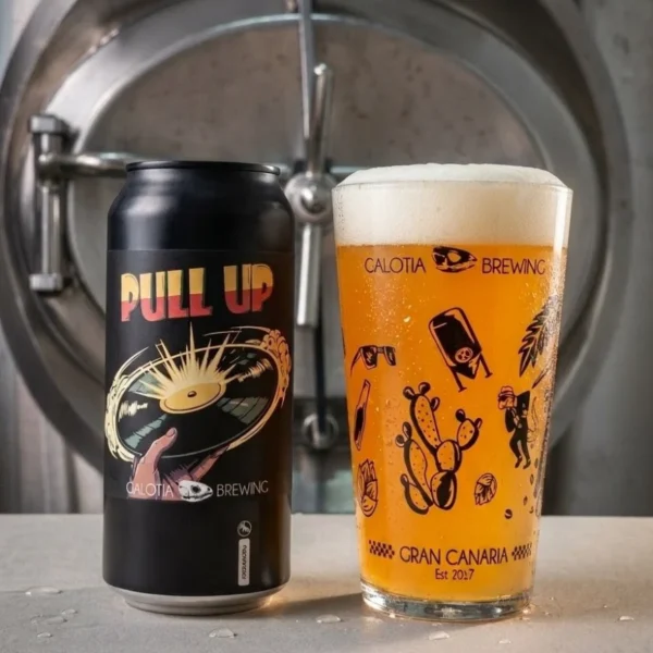 Una lata negra de cerveza Hazy IPA. el nombre es "Pull Up" de Galotia Brewing. junto a un vaso de pinta lleno de cerveza dorada con espuma, frente a un tanque de fermentación de acero inoxidable.